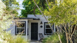 Una casa petita de color blanc amb una porta i finestres negres, envoltada de verdura i un camí cap a l'entrada.