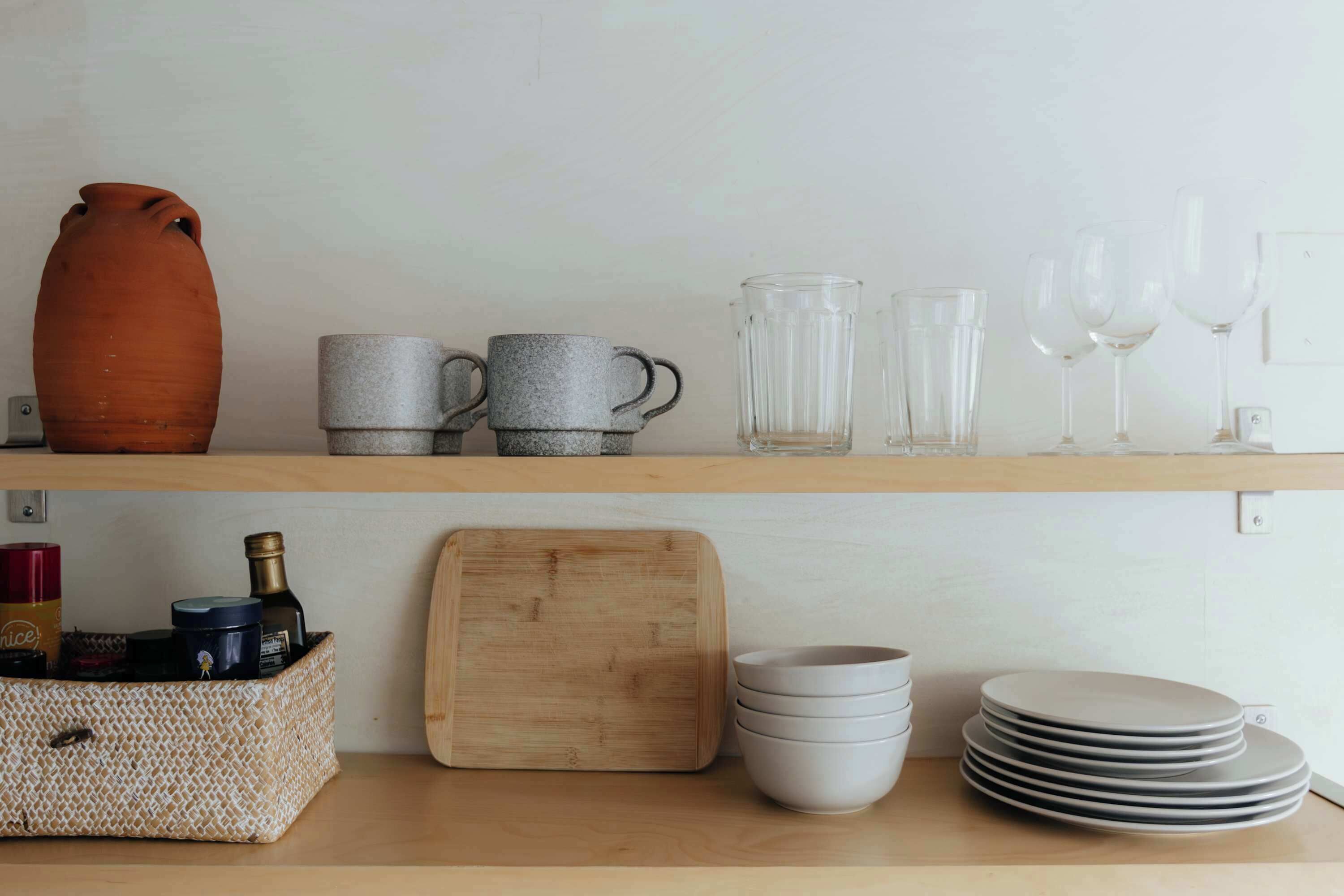 Una repisa de cocina con cerámica y cristalería variada, una tabla de cortar de madera y platos apilados.