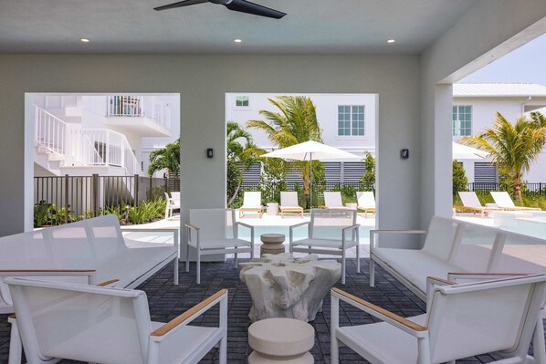 A modern outdoor seating area with white furniture, a central stone table, and a view of a pool and palm trees. - The Dreamside House at Lunara Bay (Key West)