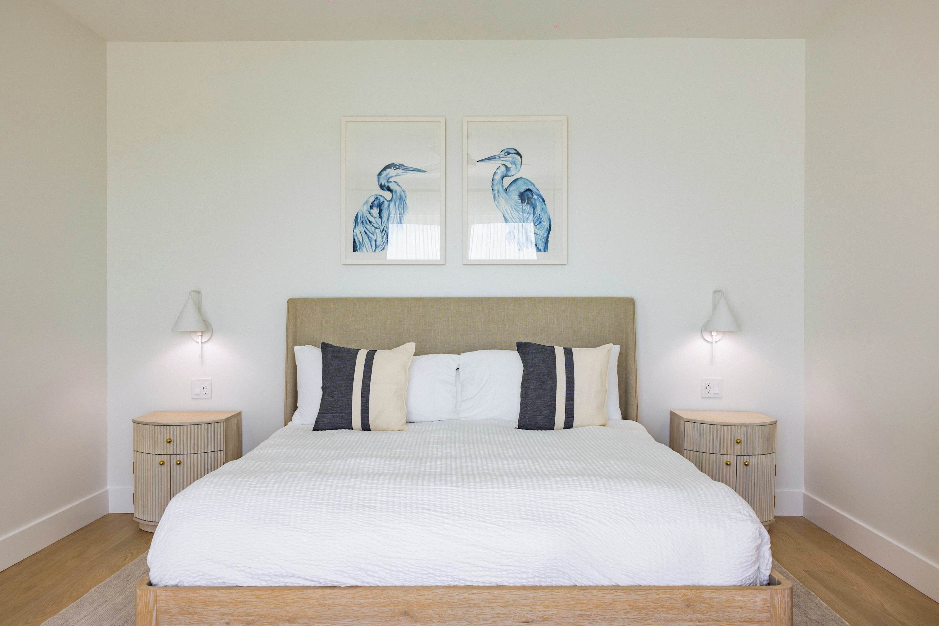 Een keurig opgemaakt bed met een houten hoofdbord, twee nachtkastjes met lampen en twee wanddecoraties van vogels.