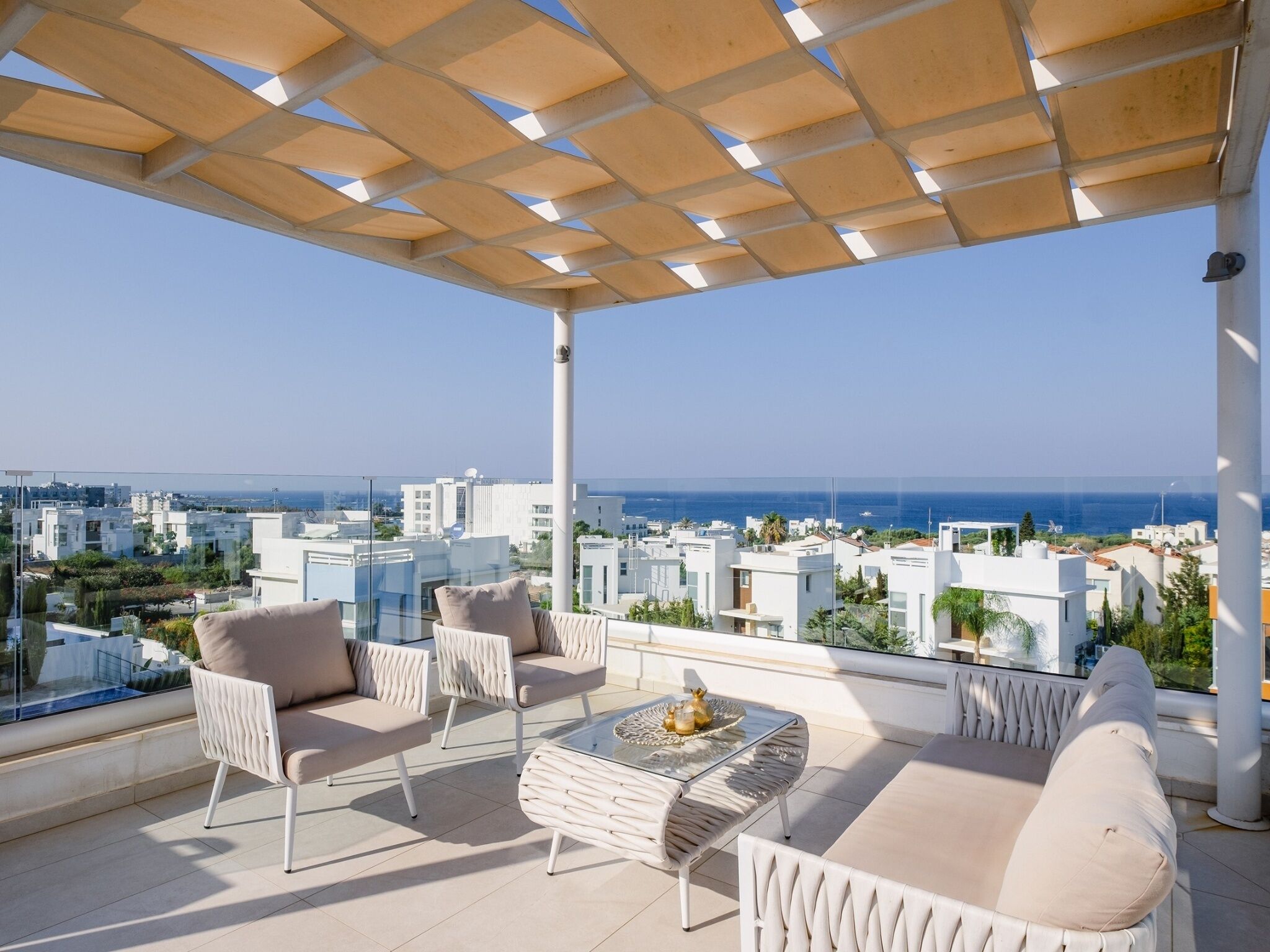 Un balcón con vistas al mar y al paisaje urbano, amueblado con sillas blancas y una mesa de vidrio.