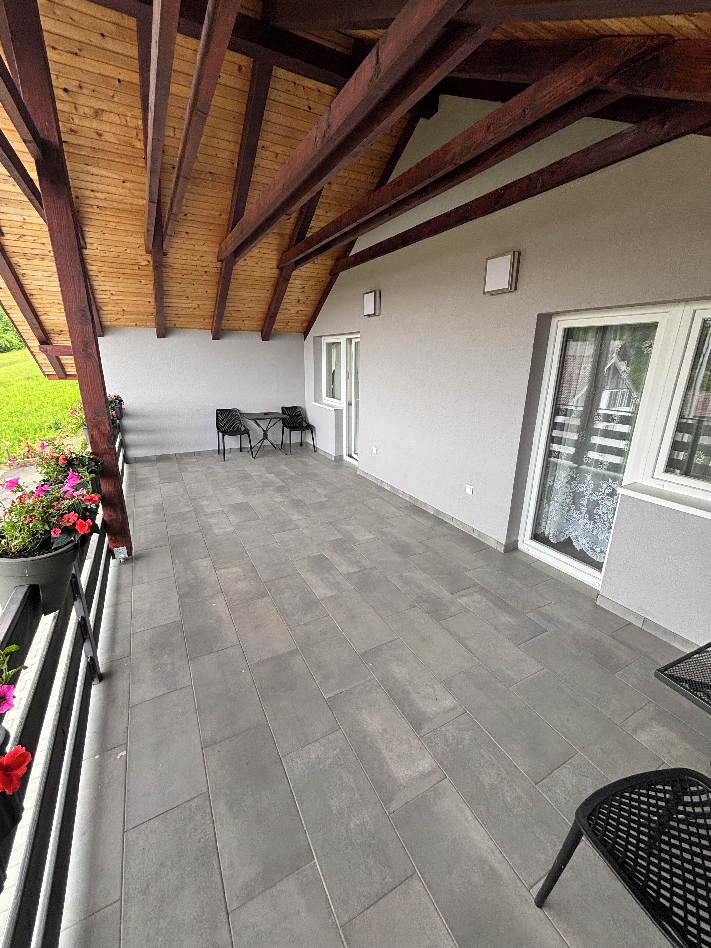 Un patio techado con suelo de baldosas, vigas de madera, una mesa con sillas y una ventana con cortinas de encaje.
