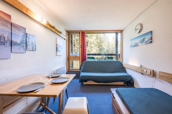 Une petite pièce comprenant un lit, une table avec des assiettes et des verres, un banc et une fenêtre donnant sur des arbres.