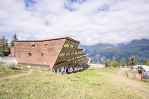 A modern building with a sloped roof, surrounded by a grassy area and mountains in the background.