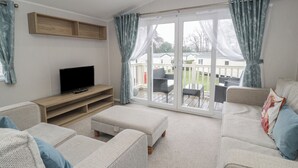 A living room with a flat-screen TV, a wooden entertainment unit, a sofa, an ottoman, and a sliding glass door leading to an outdoor patio. - Woodlands Platinum (Ruthin)