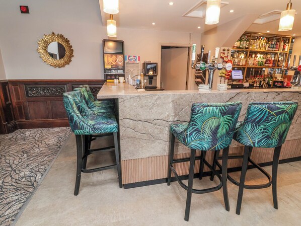 A bar area with a marble countertop, bar stools with tropical leaf upholstery, a mirror with a gold frame, and a shelf with various bottles.