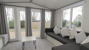 Un salon moderne avec un canapé gris, une petite table blanche et de grandes fenêtres donnant sur l’extérieur.