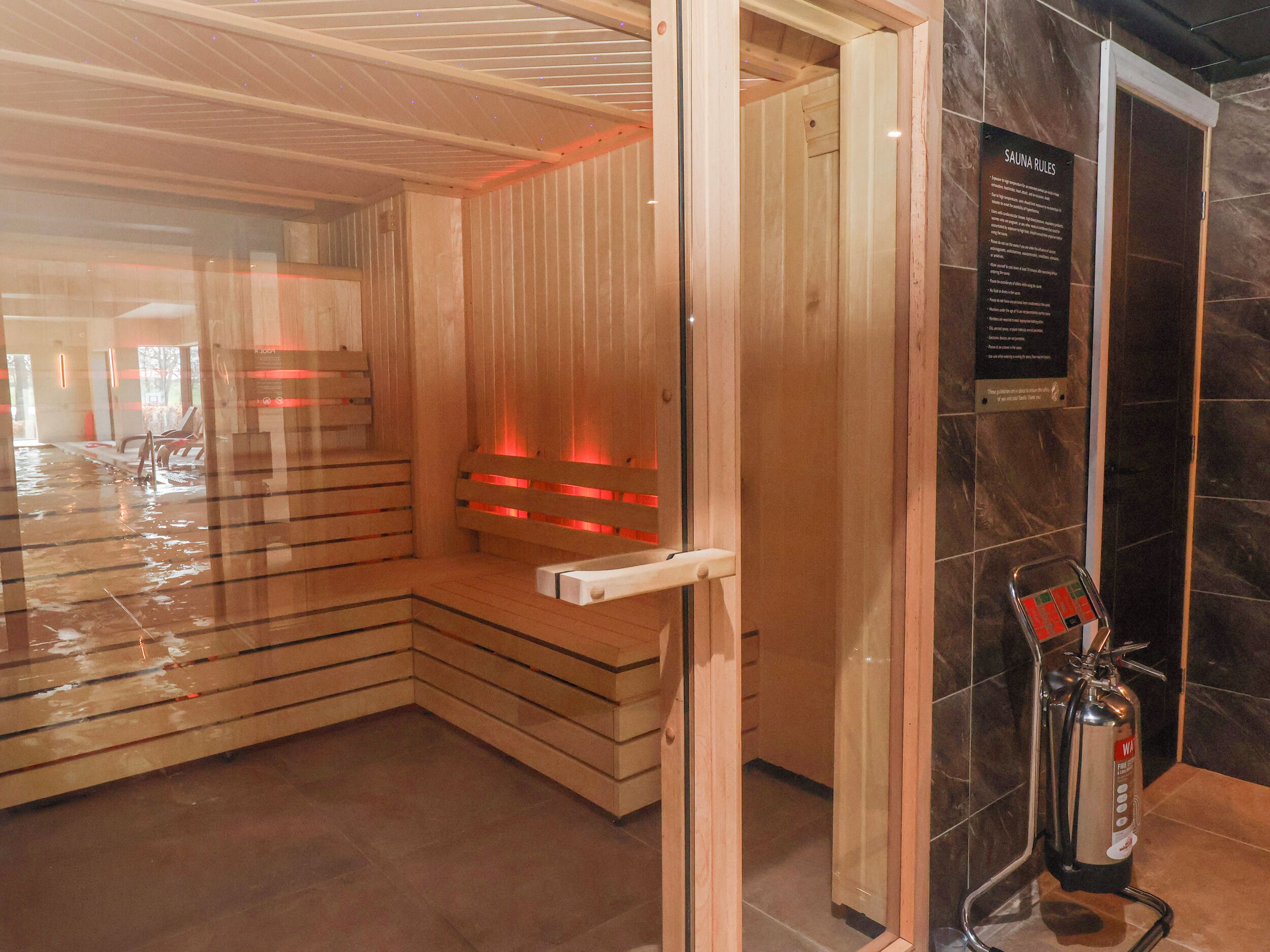 A sauna room with wooden benches, a glass door, and a fire extinguisher.