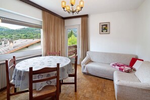 A room with a dining table, chairs, a sofa, a chandelier, a window with curtains, and a view of a green landscape.