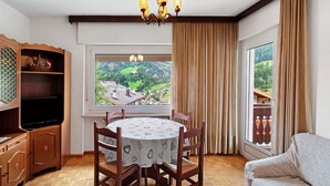 Uma sala de jantar com uma mesa redonda, cadeiras, um candelabro, uma televisão e vista para uma aldeia através de uma janela.