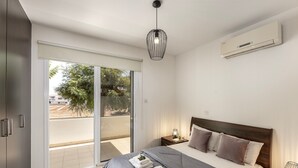 A modern bedroom with a large bed, a sliding glass door to a balcony, a ceiling light, and an air conditioning unit.