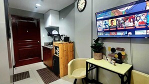 Une petite pièce avec un téléviseur diffusant différentes émissions, un meuble en bois, une horloge et une petite table sur laquelle est posé un panneau indiquant « BIENVENUE ».