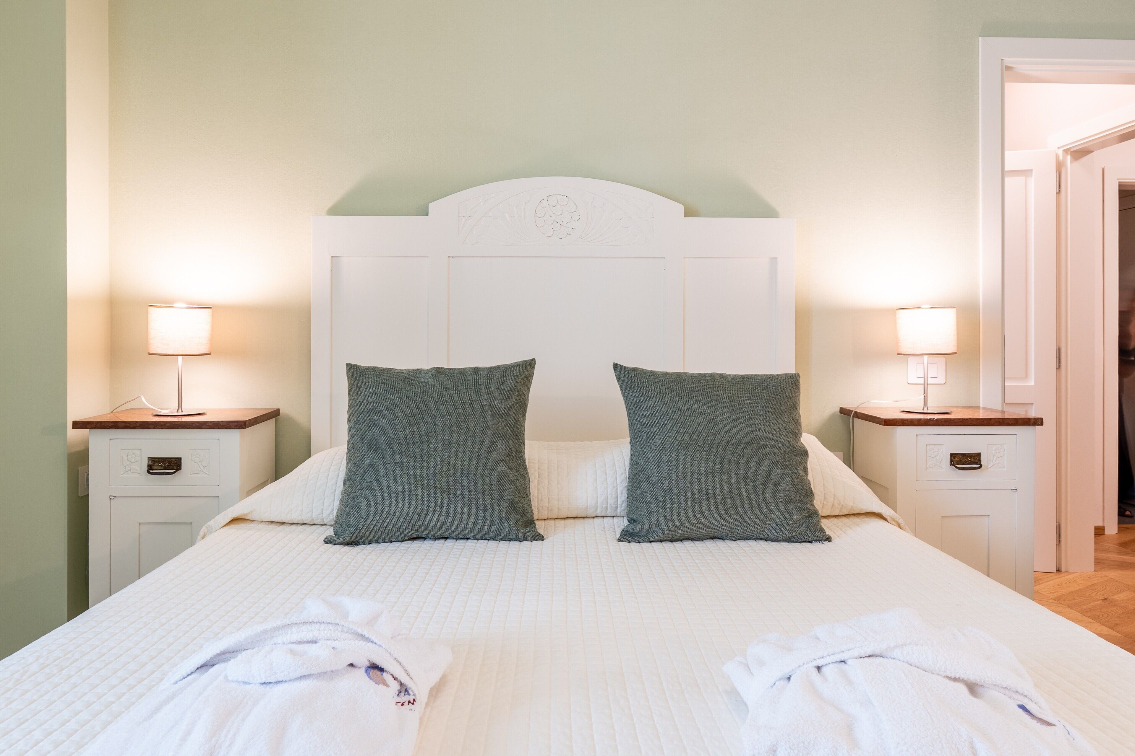 A double bed with two pillows, a bedside table with a lamp, and a door leading to another room.