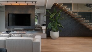 A modern living room with a large flat-screen TV, a grey sectional sofa, a large potted plant, and a staircase with wooden steps and metal railings.