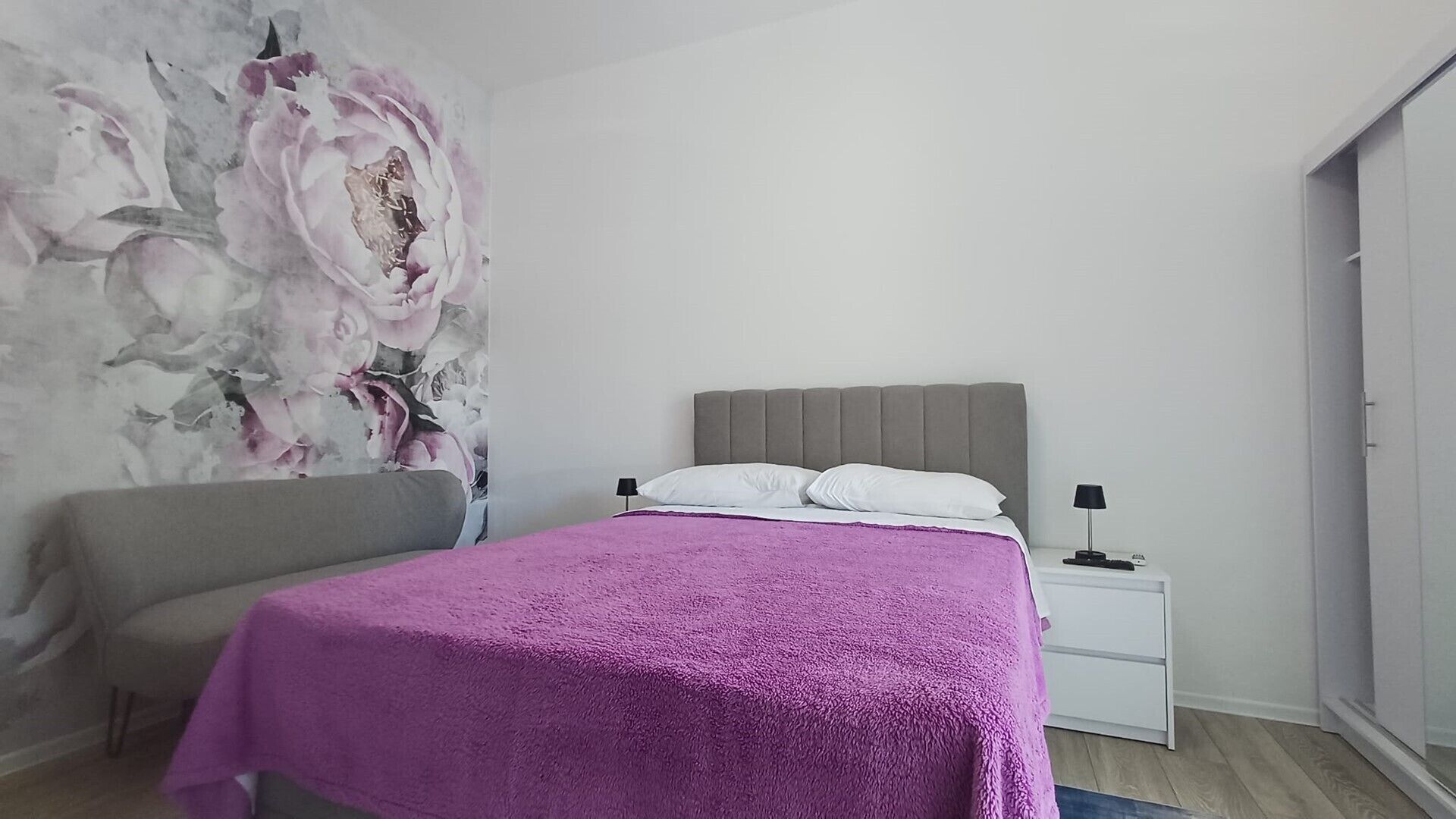 A bedroom with a bed, a nightstand, a chair, and a wall with a floral painting.