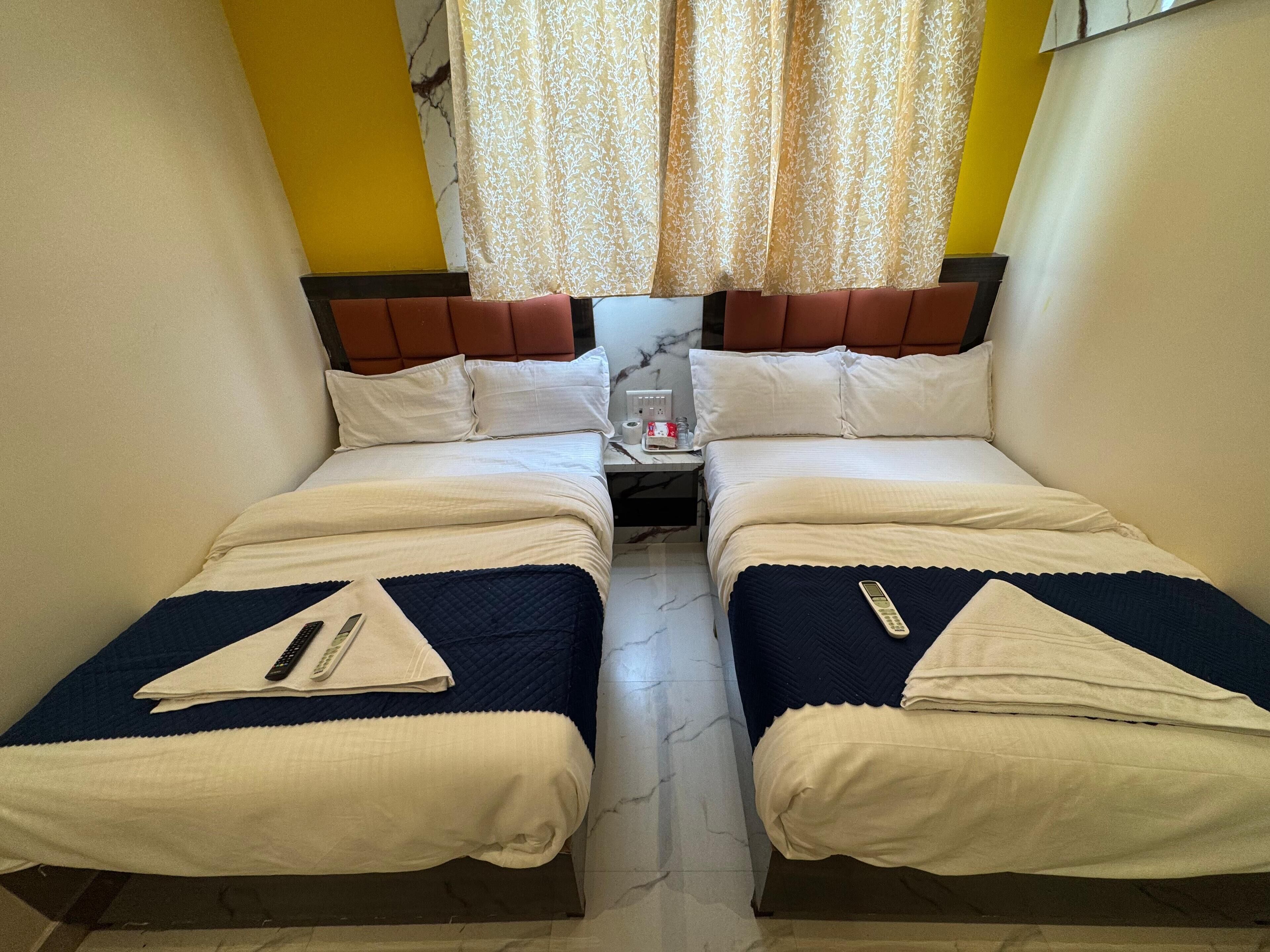 A hotel room with two beds, each with a white and blue bedspread, a nightstand between them, and a curtain with a floral pattern.