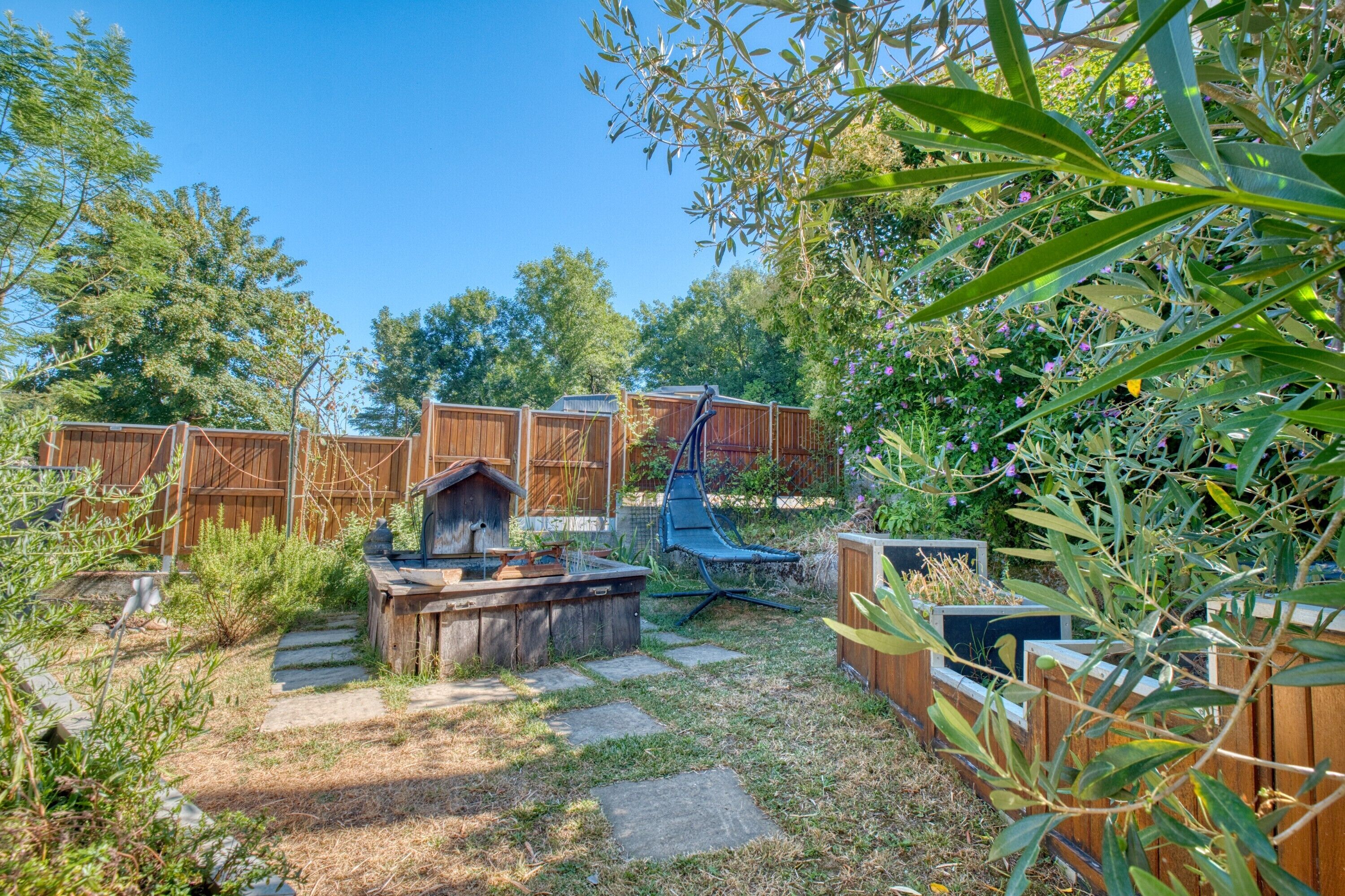 Un jardin avec une terrasse en bois, un nichoir, un foyer et diverses plantes.