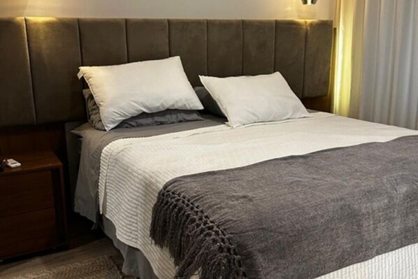 Een net opgemaakt bed met een grijze deken en twee witte kussens, een houten nachtkastje met een afstandsbediening, en een hoofdbord met ingebouwde verlichting.