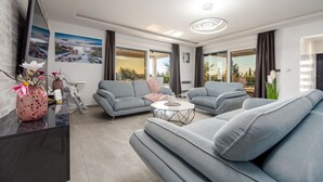 Ein modernes Wohnzimmer mit grauen Sofas, einem Couchtisch und einem Fernseher auf einem schwarzen Standfuß. Große Fenster mit Vorhängen, ein Gemälde an der Wand und eine Vase mit Blumen auf einem schwarzen Tisch.