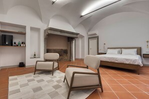A bedroom with a bed, two armchairs, a fireplace, a television, and a shelf with bottles.