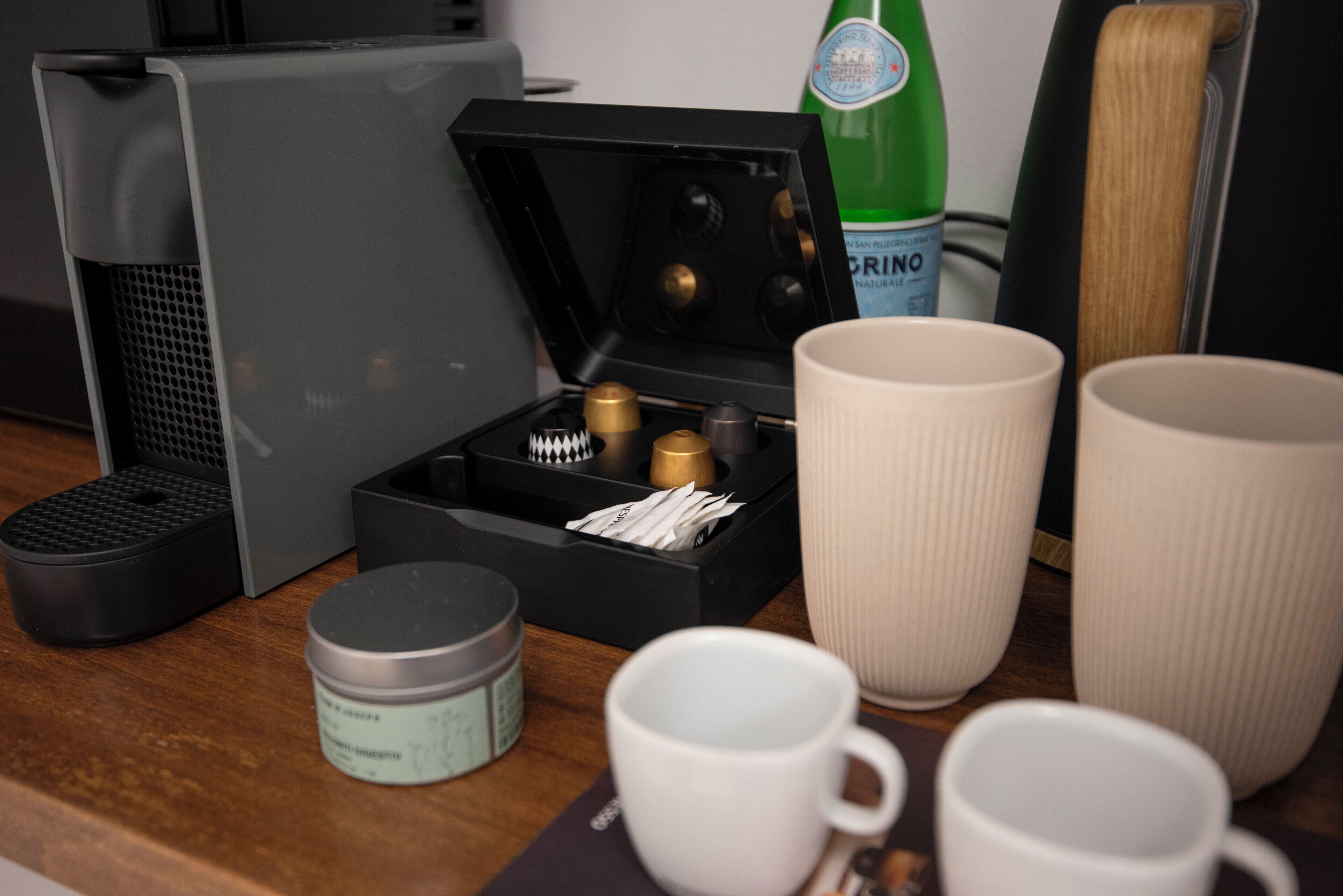 Una cafetera, una caja con cápsulas de café, una botella verde y varios tazas blancas sobre una superficie de madera.