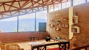 Una habitació àmplia amb un sostre alt, parets de maó i grans finestres que ofereixen vista a la muntanya. Hi ha una prestatgeria, una taula i cadires de vimet.