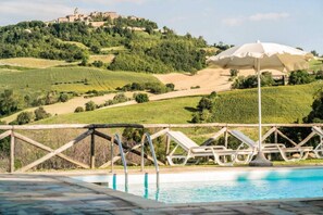 A pool area with lounge chairs and an umbrella, surrounded by terraced vineyards and a hilltop building.