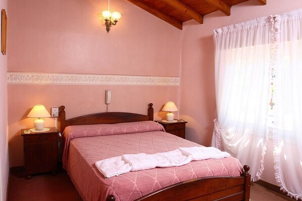 Une chambre à coucher avec un lit en bois, deux lampes de chevet, un panneau décoratif au mur et une fenêtre ornée de rideaux légers.
