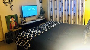 A bedroom with a bed, a television, a small table with a picture frame, and a curtain with a floral pattern. - Your home away from home. (नवी मुंबई)