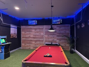 Ein Billardtisch mit roter Filzoberfläche, umgeben von einer Holzwand und einem mit grünem Teppich belegten Boden. Links befindet sich ein Billardständer und ein kleiner Tisch mit einer Spielkonsole.