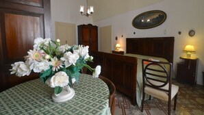 A dining area with a round table covered by a patterned tablecloth, a vase of white flowers, a wooden chair, a wooden cabinet, and a wall-mounted picture.