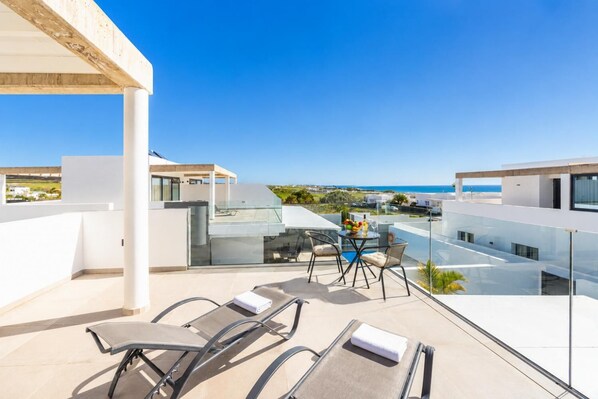 Una terraza moderna en la azotea con barandilla de cristal, mobiliario exterior y vistas al mar.