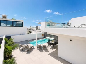 A rooftop pool area with lounge chairs, a dining table, and a glass balustrade overlooking the pool.