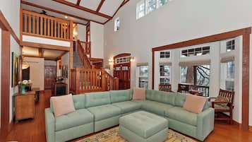 A spacious living room with a large sectional sofa, a wooden staircase, and a view of the outdoors through large windows.
