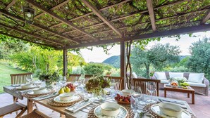 An outdoor dining area with a pergola, wooden tables, and chairs, surrounded by lush greenery and a scenic view. - Iconic Romantic Farmhouse with Pool in Lucca (Lucca)