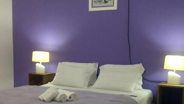 A bedroom with a purple wall, a bed with white linens, two bedside lamps, and a small framed picture on the wall. - Amber Sul Hospedagem (Gramado)