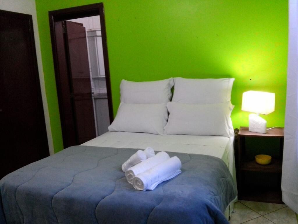 Una habitación con cama, almohadas, una mesita de noche con lámpara y una toalla sobre la cama.