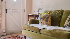 A green sofa with patterned cushions, a white door, and a small table with books. - Bertie's Cottage, East Sussex (Robertsbridge)