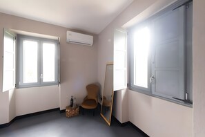 A room with a large window, an air conditioning unit, a chair, a mirror, and a small table with a bottle and glasses.
