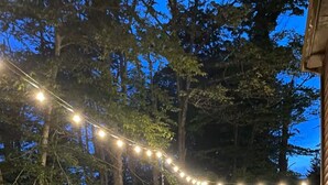 Terraza de madera con luces colgantes, rodeada de árboles.