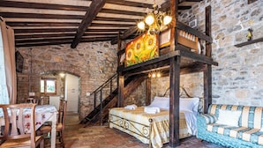 A rustic room with a stone wall, wooden beams, a bunk bed, a dining table with chairs, and a sofa.