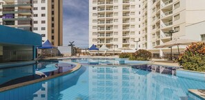 A swimming pool with a curved design, surrounded by lounge chairs and umbrellas, with high-rise buildings in the background. - Apartamento Caldas Novas com Piscinas Termais (Caldas Novas)
