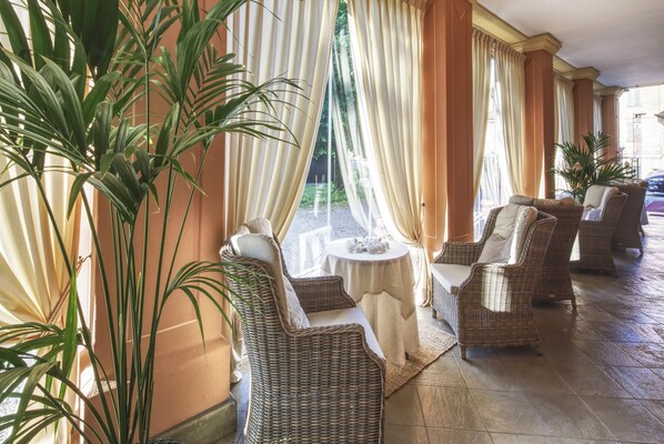 A covered outdoor seating area with wicker furniture and a small table, surrounded by large windows with curtains.