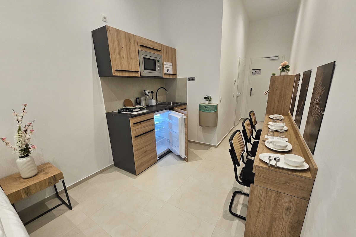 A compact kitchen with modern appliances, a dining area with chairs and a table set for four, and a small side table with a vase.