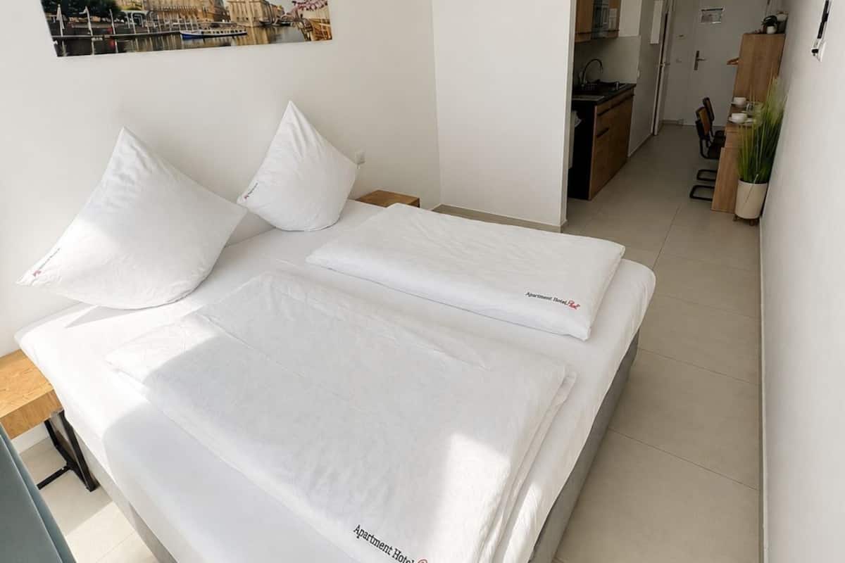 A modern hotel room with a large bed, a small kitchenette, and a cityscape painting on the wall.
