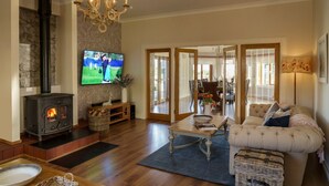 A cozy living room with a fireplace, a television, a sofa, and a dining area visible through glass doors. - St Andrews Retreat - Ten Bedroom Villa, Sleeps 20 (Cupar)