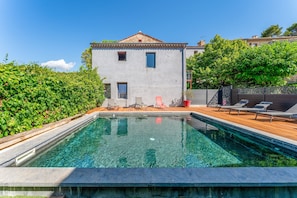 A swimming pool with a wooden deck, a building with a tiled roof, and a hedge. - Apartment 'Côté Vigne' with Shared Pool, Private Terrace and Wi-Fi (Rasteau)