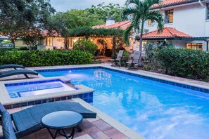 Une piscine avec des chaises longues et un jacuzzi, entourée de verdure, et une maison avec un toit recouvert de tuiles rouges.