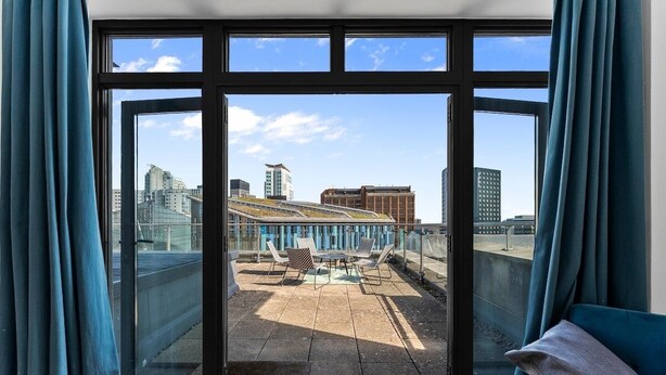Une pièce dotée de grandes fenêtres donnant sur un paysage urbain, avec des bâtiments et une terrasse sur le toit aménagée avec du mobilier d’extérieur.
