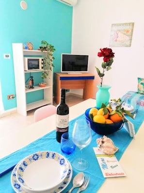 Uma área de refeições com uma mesa posta para dois, uma garrafa de vinho, uma cesta de frutas e um vaso com uma flor.